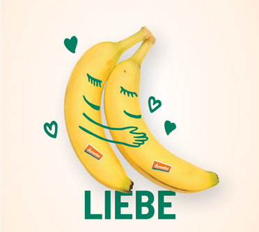 Zwei Bananen umarmen sich, mit gemalten Gesichtern, Händen und Herzen und der Aufschrift Liebe 